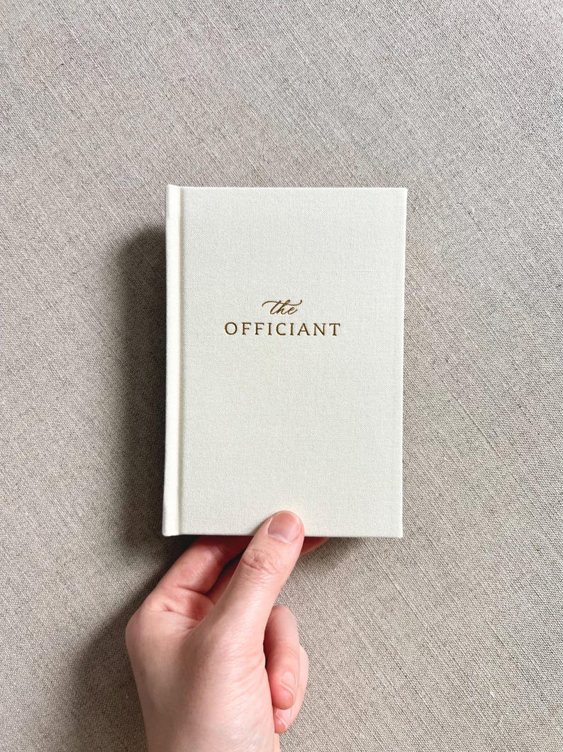 May include: A small, off-white book with the words "the Officiant" in gold lettering. The book is held by a hand against a neutral-toned fabric background. The book's cover has a textured appearance.