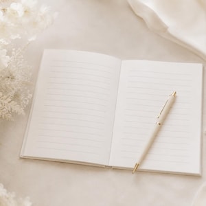 May include: An open notebook with white lined pages lies flat, accompanied by a cream-colored ballpoint pen with gold-tone accents resting diagonally across the right page. The notebook features a clean, minimalist design with crisp white paper.