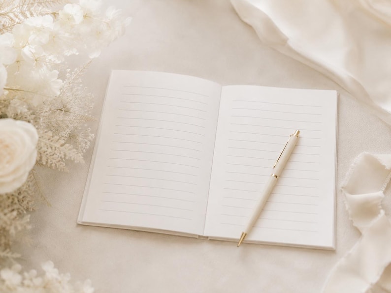 May include: An open notebook with white lined pages lies flat, accompanied by a cream-colored ballpoint pen with gold-tone accents resting diagonally across the right page. The notebook features a clean, minimalist design with crisp white paper.