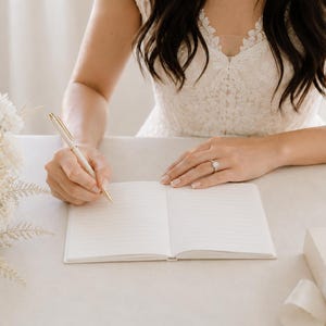 May include: An open, lined notebook with white pages resting on a flat surface. A gold-toned pen is held in position to write on the paper. The notebook is positioned near a bouquet of white flowers and a gift box with a white ribbon.