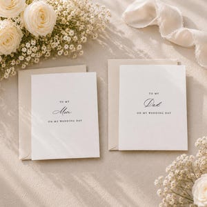 May include: Two white greeting cards with matching envelopes, each featuring black serif and script text. One card reads TO MY Mom ON MY WEDDING DAY, and the other reads TO MY Dad ON MY WEDDING DAY. The cards are displayed with white roses and baby's breath.