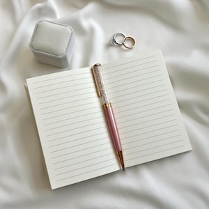 May include: An open notebook with cream-colored lined pages, a pink ballpoint pen with a crystal-filled barrel, a light gray velvet ring box, and two metallic rings, one featuring a clear stone, resting on a soft white fabric surface.
