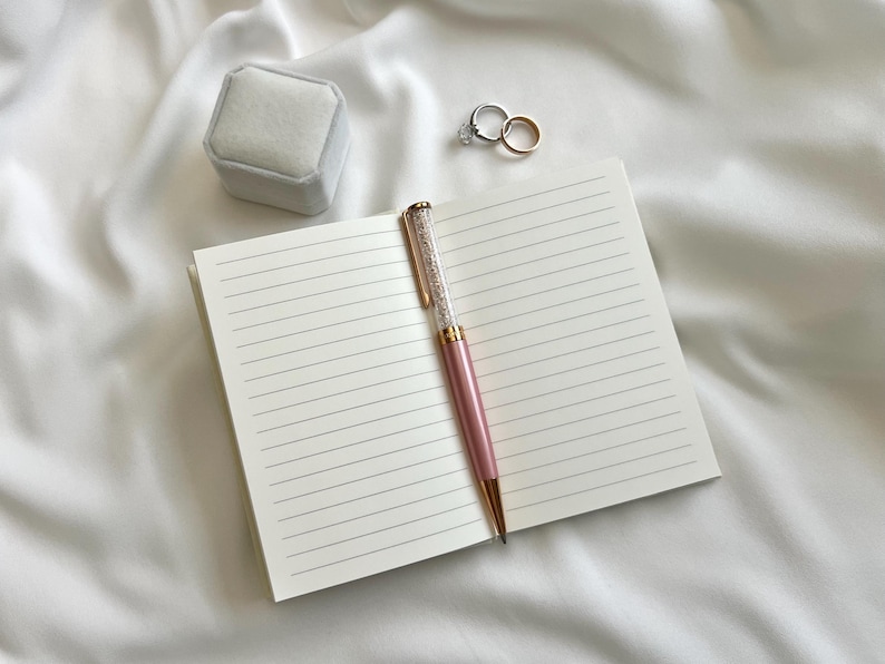 May include: An open notebook with cream-colored lined pages, a pink ballpoint pen with a crystal-filled barrel, a light gray velvet ring box, and two metallic rings, one featuring a clear stone, resting on a soft white fabric surface.
