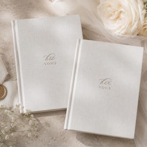 May include: Two white hardcover vow books with textured fabric covers. One book features the text his vows in elegant script, while the other features the text her vows in the same script. Both books are rectangular with a clean, simple design.