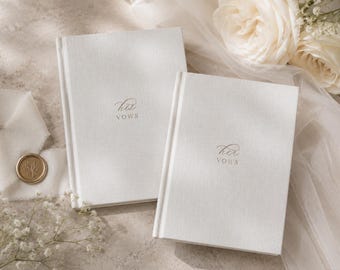 May include: Two white hardback vow books with textured fabric covers. One book features the text his vows in elegant script, while the other features the text her vows in the same script. Both books are rectangular with a clean, simple design.
