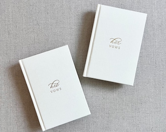 May include: Two white vow books with gold lettering that reads "Her Vows" and "His Vows". The books are rectangular with a simple, elegant design. The background is a neutral gray fabric.