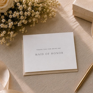 May include: A white rectangular greeting card with black serif text that reads Thank you for being my Maid of Honor. The card is placed next to a gold pen, a beige gift box with a satin ribbon, and a bouquet of cream roses and white baby's breath.