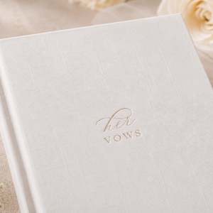 May include: A white hardcover book with a textured fabric cover. The front cover features the embossed text "her VOWS" in a combination of elegant script and clean sans-serif lettering centered on the page. The book has a clean, minimalist design.