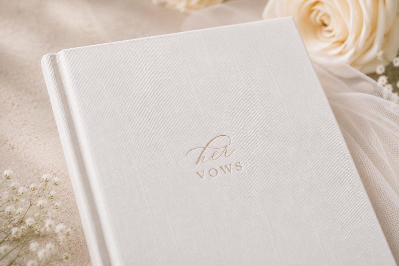 May include: A white hardcover book with a textured fabric cover. The front cover features the embossed text "her VOWS" in a combination of elegant script and clean sans-serif lettering centered on the page. The book has a clean, minimalist design.