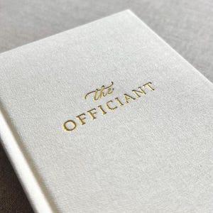 May include: A close-up of a white book with the gold-colored text "the OFFICIANT" on the cover. The book has a textured, linen-like appearance. The background is a soft, neutral color.