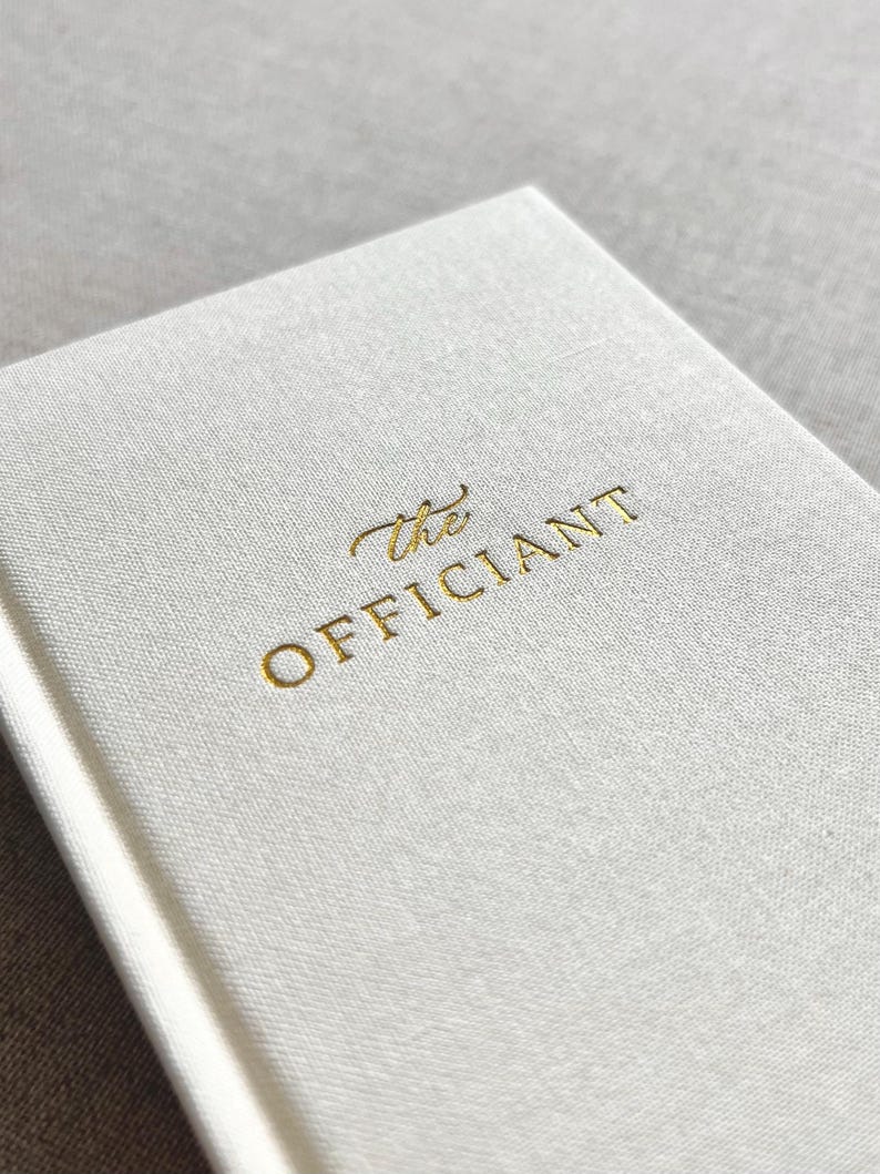 May include: A close-up of a white book with the gold-colored text "the OFFICIANT" on the cover. The book has a textured, linen-like appearance. The background is a soft, neutral color.