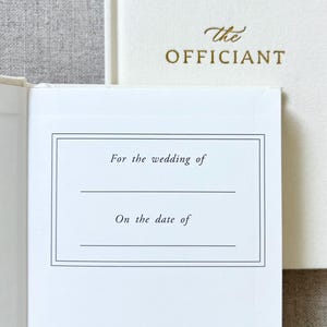 May include: Off-white wedding officiant book with gold lettering. The open page has a bordered section with the text "For the wedding of" and "On the date of". The cover of the book on the right has the word "the" and "OFFICIANT" in gold.