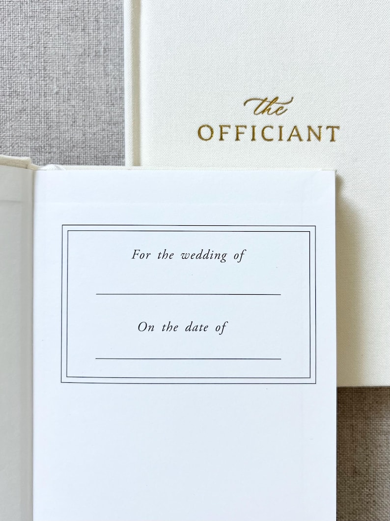 May include: Off-white wedding officiant book with gold lettering. The open page has a bordered section with the text "For the wedding of" and "On the date of". The cover of the book on the right has the word "the" and "OFFICIANT" in gold.