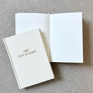May include: Ivory-colored notebook with the gold text "The Officiant" on the cover, next to an open notebook with lined pages. The notebooks are on a gray textured surface. The open notebook displays several lined pages.