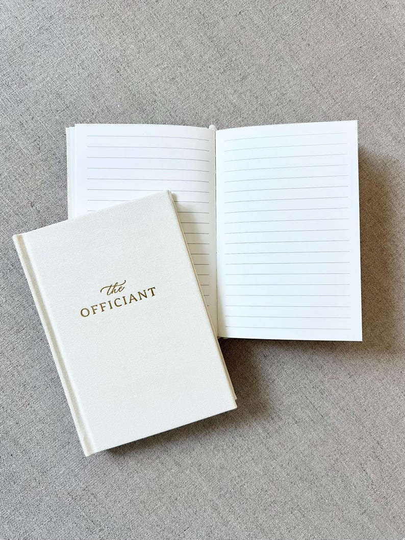 May include: Ivory-colored notebook with the gold text "The Officiant" on the cover, next to an open notebook with lined pages. The notebooks are on a gray textured surface. The open notebook displays several lined pages.
