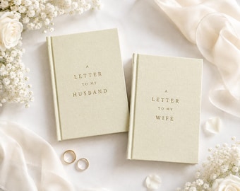 May include: Two light beige hardcover books with gold embossed text. One book reads A LETTER TO MY HUSBAND and the other reads A LETTER TO MY WIFE. Two gold wedding bands are positioned near the books, surrounded by white floral accents.