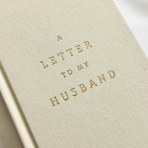 May include: A cream-colored fabric-bound journal featuring the gold-embossed text A LETTER TO MY HUSBAND centered on the front cover. The book has a textured linen-like finish and a clean, simple design with a vertical spine visible on the left side.