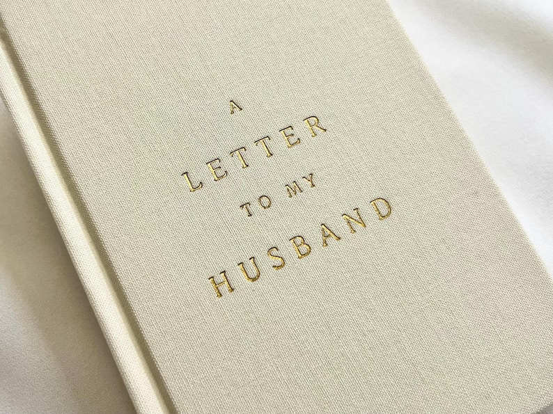 May include: A cream-colored fabric-bound journal featuring the gold-embossed text A LETTER TO MY HUSBAND centered on the front cover. The book has a textured linen-like finish and a clean, simple design with a vertical spine visible on the left side.