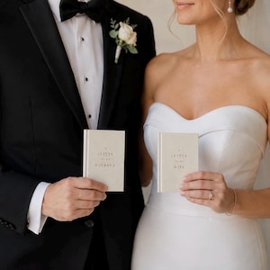 May include: Two small, off-white hardcover books with gold foil lettering. One book reads A LETTER TO MY HUSBAND, and the other reads A LETTER TO MY WIFE. The books are held in front of a black tuxedo jacket and a white strapless wedding dress.