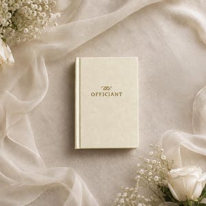 May include: A cream-colored hardcover book with the words "the" and "OFFICIANT" embossed in gold lettering on the front cover. The book is surrounded by white roses and delicate baby's breath flowers arranged on a soft, flowing fabric.
