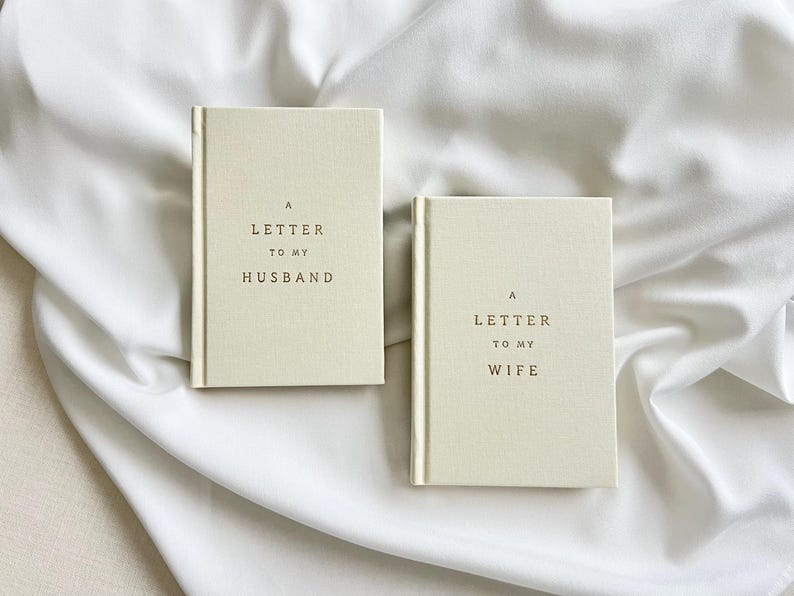 May include: Two cream-colored hardcover books with a fabric-like texture. The left book features the text A LETTER TO MY HUSBAND in dark brown lettering. The right book features the text A LETTER TO MY WIFE in dark brown lettering.