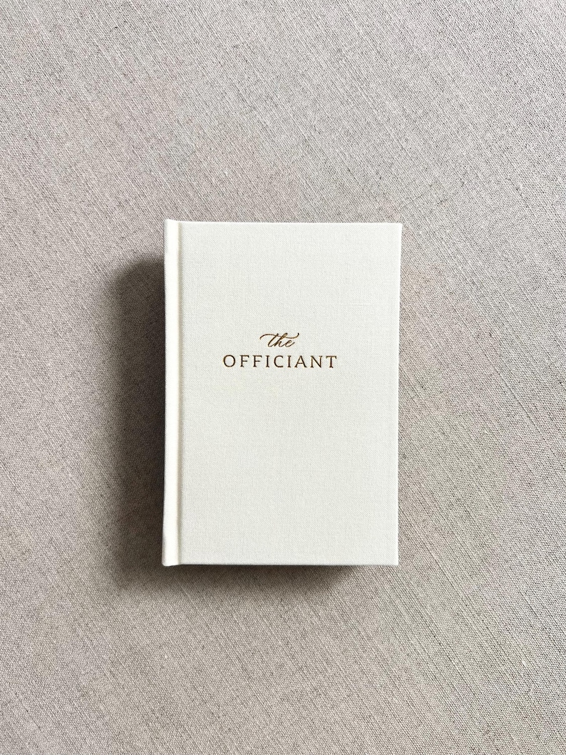 May include: A small, off-white book with the words "the officiant" in gold lettering on the cover. The book has a textured, linen-like appearance and a clean, minimalist design. The book is likely a wedding planning or ceremony guide.