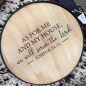 May include: Round wooden serving tray with a black metal rim and handles. The tray features the text "AS FOR ME AND MY HOUSE, we will serve the Lord. - JOSHUA 24:15" in black lettering. The tray has a light wood grain.