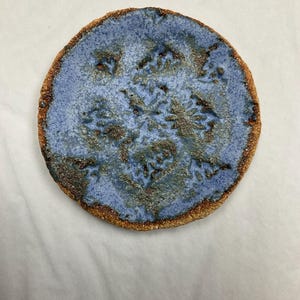 May include: A small, round ceramic plate with a blue glaze and brown speckles. The plate has a unique, organic design that resembles a flower or a tree.