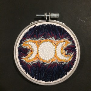 May include: A circular embroidery hoop with a white fabric background and a triple moon symbol embroidered in yellow, orange, and white thread. The background is filled with a design of blue, red, and white thread.