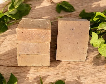 May include: Four rectangular bars of soap are stacked, with another bar beside them. The soap is a light brown color with small dark specks. Green leaves are scattered around the soap on a wooden surface, creating a natural aesthetic.