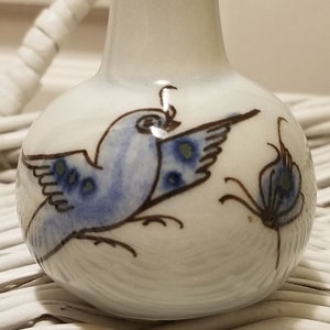 May include: A small white ceramic vase with a blue and brown bird design. The vase has a narrow neck and a round body.