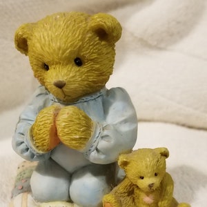 May include: A yellow teddy bear wearing a blue sleeper is kneeling on a colorful patchwork quilt. A smaller yellow teddy bear is sitting next to the larger bear.