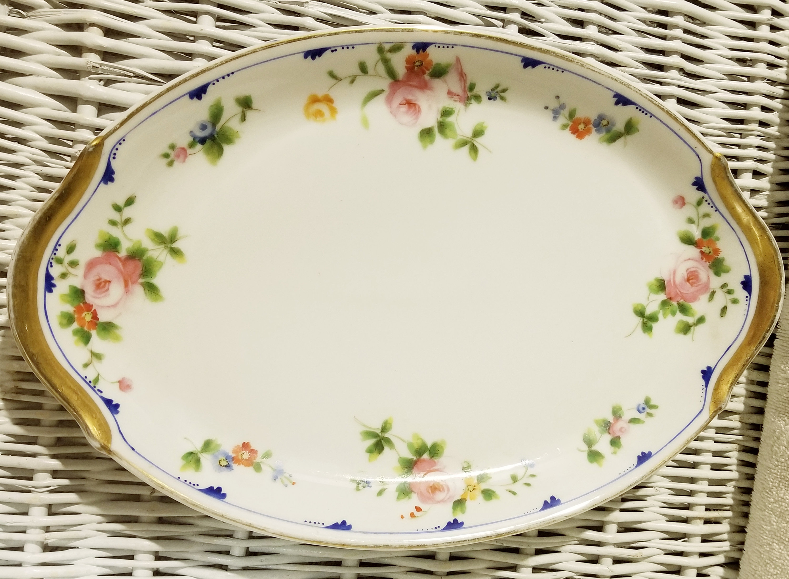 May include: An oval serving platter with a white background and a gold rim. The platter is decorated with a floral pattern in shades of pink, blue, yellow, and green.