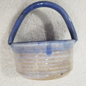 May include: A small, white ceramic planter with a blue glazed handle. The planter has a light brown glaze with white streaks and a blue glaze around the rim.