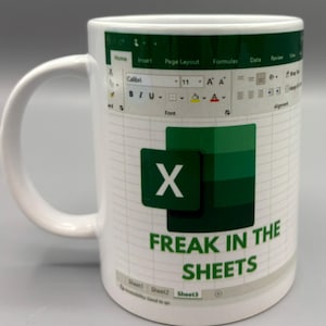 May include: White ceramic mug featuring an Excel spreadsheet design with the Microsoft Excel logo and the text "FREAK IN THE SHEETS" in green. The mug has a white handle and a grid pattern background.