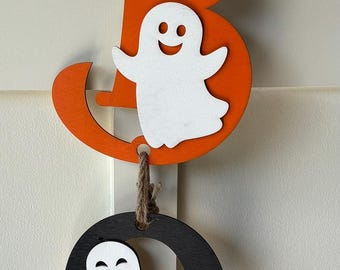May include: A Halloween-themed wooden hanging decoration. The decoration features three circular and letter-shaped cutouts with ghost and bat designs in orange, black, and purple. The pieces are connected by twine.