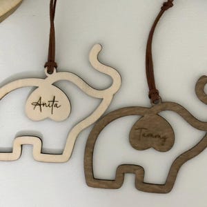 Op de afbeelding: Twee houten olifantenornamenten, &eacute;&eacute;n licht en &eacute;&eacute;n donker, met bruine ophangkoorden. Elke olifant heeft een hartvormige uitsparing met een naam: "Anita" en "Tammy". De ornamenten zijn waarschijnlijk voor decoratie of personalisatie.