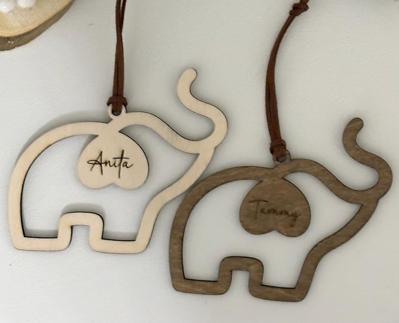 Op de afbeelding: Twee houten olifantenornamenten, &eacute;&eacute;n licht en &eacute;&eacute;n donker, met bruine ophangkoorden. Elke olifant heeft een hartvormige uitsparing met een naam: "Anita" en "Tammy". De ornamenten zijn waarschijnlijk voor decoratie of personalisatie.