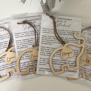 Op de afbeelding: Houten boekenleggers in de vorm van een olifant met gegraveerde namen, elk bevestigd aan een bruin koord. De boekenleggers zijn verpakt in transparant plastic met tekst over zusterschap. De namen zijn Lauren, Came, Morgan en Emily.