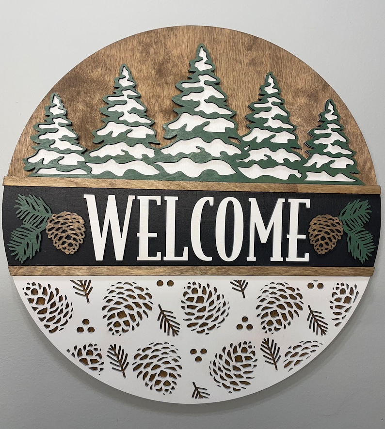May include: A circular wooden welcome sign with a woodland theme. The top section has a brown background with green and white trees. The word "WELCOME" is in white on a black background. The bottom section has a white background with pine cone cutouts.