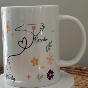 Pode incluir: Caneca de cer&acirc;mica branca decorada com um padr&atilde;o floral e contornos de estados. A caneca apresenta os nomes "Wendy" e "Brenda" com uma linha em forma de cora&ccedil;&atilde;o conectando a Fl&oacute;rida e a Carolina do Norte. Acentos florais e estrelas adicionais.