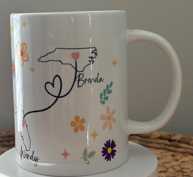Pode incluir: Caneca de cer&acirc;mica branca decorada com um padr&atilde;o floral e contornos de estados. A caneca apresenta os nomes "Wendy" e "Brenda" com uma linha em forma de cora&ccedil;&atilde;o conectando a Fl&oacute;rida e a Carolina do Norte. Acentos florais e estrelas adicionais.