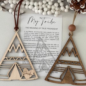 May include: Two wooden triangle ornaments with mountain and teepee designs. One is light-colored, the other is dark. Both have a leather cord for hanging. The light ornament has a heart with the name "Sandra" on it. A card reads "My Tribe".