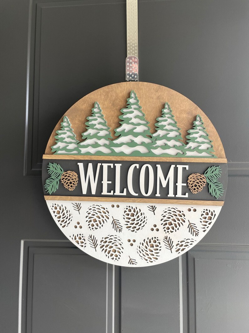 May include: A circular wooden welcome sign with a forest theme. It features a light brown background with green and white trees, a grey band with the word "WELCOME", and a white section with pine cone cutouts. A silver ribbon is attached for hanging.