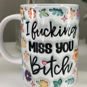 Pode incluir: Caneca de cer&acirc;mica branca com o texto "I fucking MISS YOU Bitch" em escrita preta. A caneca &eacute; decorada com desenhos florais e de folhas coloridas. A caneca tem uma al&ccedil;a branca.