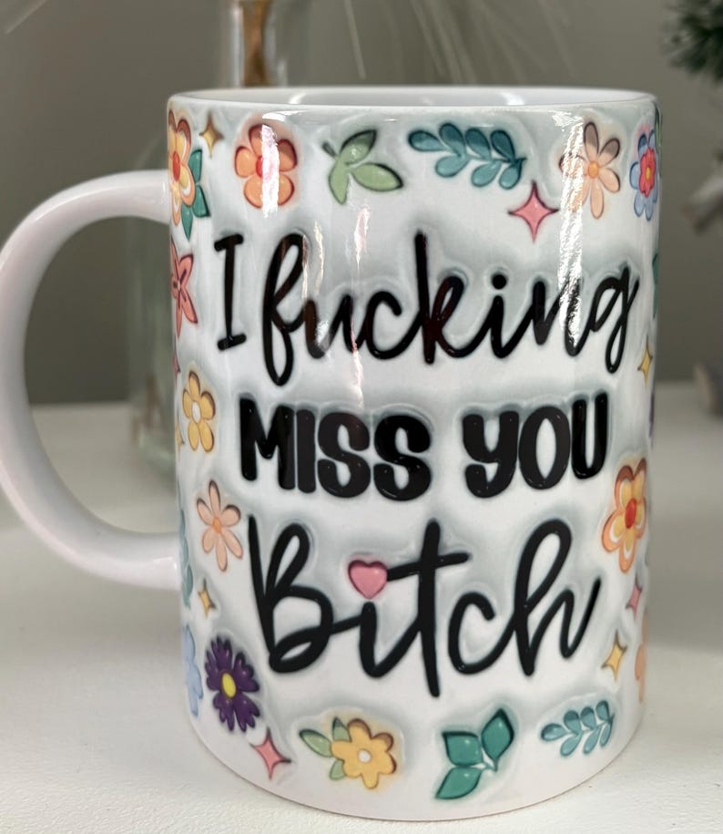 Pode incluir: Caneca de cer&acirc;mica branca com o texto "I fucking MISS YOU Bitch" em escrita preta. A caneca &eacute; decorada com desenhos florais e de folhas coloridas. A caneca tem uma al&ccedil;a branca.