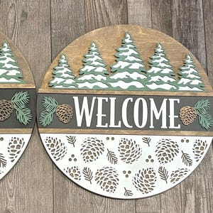 May include: Three circular wooden signs with a forest theme. Each sign features a row of green and white trees, the word "WELCOME" in white, and a pine cone design. The bottom half has a white pine cone pattern.