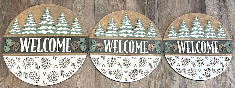 May include: Three circular wooden signs with a forest theme. Each sign features a row of green and white trees, the word "WELCOME" in white, and a pine cone design. The bottom half has a white pine cone pattern.