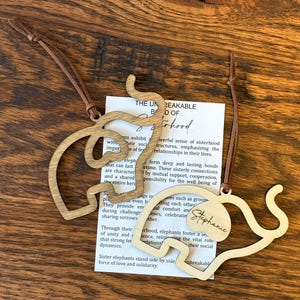 Op de afbeelding: Twee houten olifantvormige ornamenten met bruine leren koorden. E&eacute;n ornament is gegraveerd met de naam "Stephanie". Een witte kaart met tekst over zusterschap is zichtbaar op de achtergrond. De ornamenten liggen op een houten oppervlak.