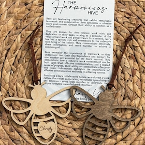 May include: Two wooden bee-shaped ornaments with leather cords. One is light-colored, the other is stained a darker brown. The light-colored bee has a heart cutout with the name "Christy" inside. The ornaments are displayed on a woven background.